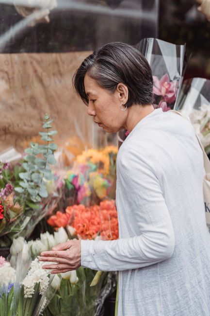 A woman with short dark hair, dressed in a light grey blazer, is selecting flowers at an outdoor floral display. She is gently touching a cluster of white tulips with her right hand while facing sidew