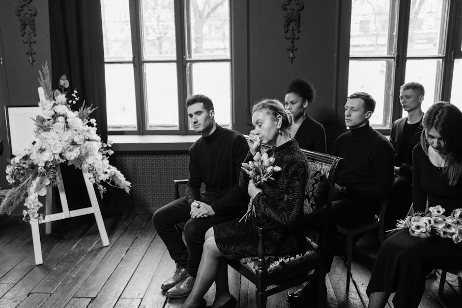 A black and white photograph showing a group of six people seated in a somber setting, likely attending a funeral or memorial service. The attendees are dressed in dark, formal clothing, with some hol