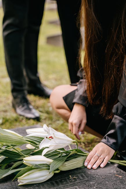 A person arranging a bouquet of white lilies with large green leaves on a black stone surface outdoors, surrounded by other individuals dressed in dark clothing. The lilies have open petals revealing 