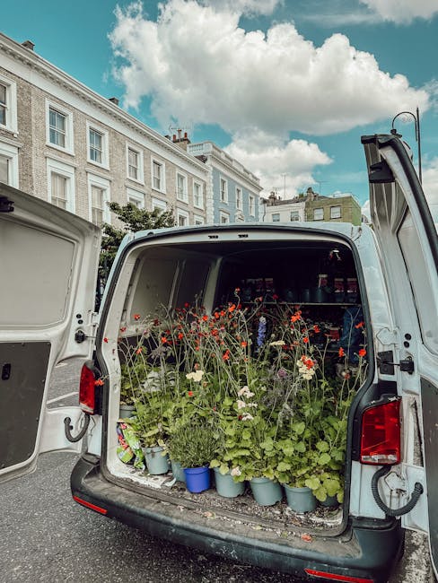 The open rear of a white delivery van filled with a variety of fresh flowers and foliage, including red poppies, white daisies, and green leafy stems, arranged in light blue pots and scattered loosely
