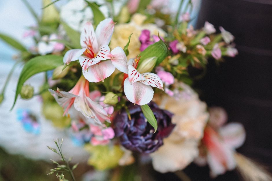 A close-up view of a floral bouquet featuring pink and white alabaster lilies with streaks of red, dark purple lisianthus, and soft pink snapdragons, all arranged with green foliage. The flowers are d