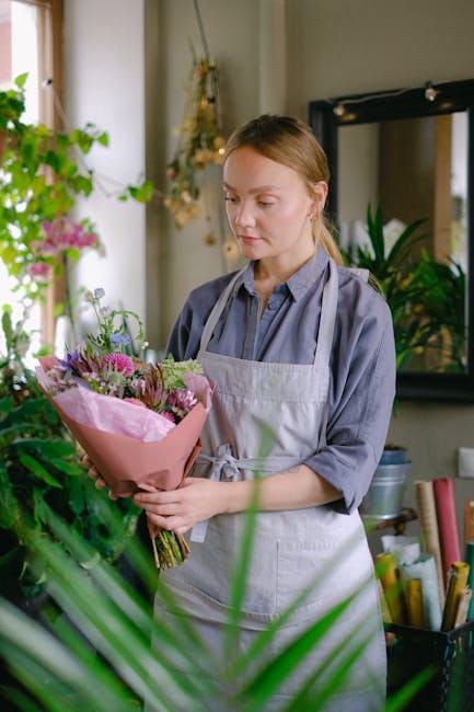 A young woman with fair skin and light brown hair tied back, dressed in a light grey apron over a blue shirt, is holding a bouquet of fresh flowers wrapped in pink paper. The bouquet features a variet
