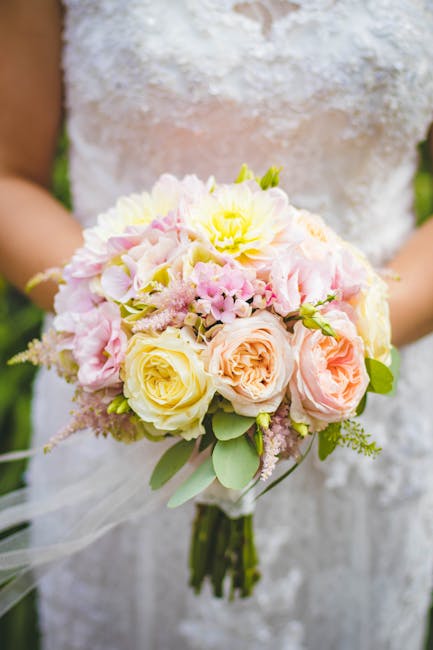 A bridal bouquet featuring a combination of soft pastel roses in shades of pale yellow, peach, and blush pink, accented with clusters of light pink and white hydrangeas, along with delicate greenery a