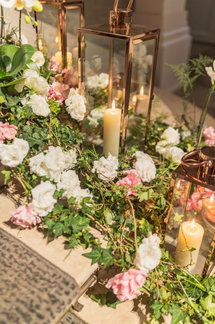A floral arrangement featuring a combination of white and soft pink flowers, including roses and peonies, complemented by lush green foliage, arranged along a table. The display includes three elegant